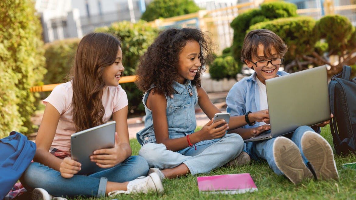 Three children laughing together on the grass with a tablet, a phone, and a laptop